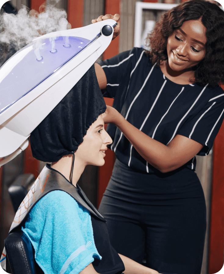 A stylist smiling at a client under a hair steamer